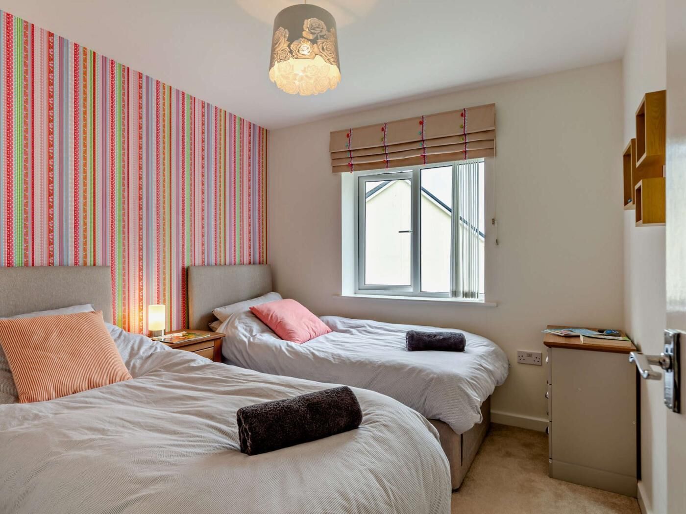 Une chambre avec deux lits, une fenêtre avec des stores, une table de chevet avec une lampe et un mur recouvert d’un papier peint à rayures.