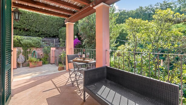 Eine überdachte Terrasse im Freien mit Tisch und Stühlen, einer Korbsitzbank und Blick auf üppiges Grün.