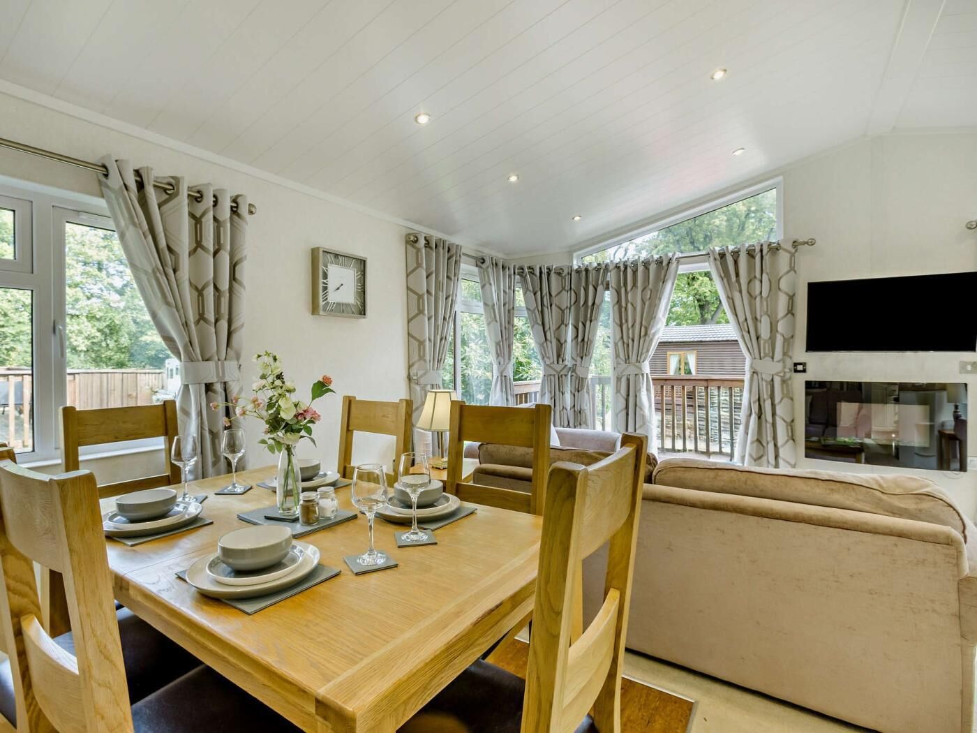 Un coin repas avec une table en bois dressée pour quatre personnes, des chaises, un vase avec des fleurs et un canapé en arrière-plan.