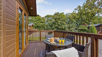 Une terrasse en bois avec une table ronde et des chaises en osier, une porte vitrée donnant sur un espace abrité, et une vue sur la verdure environnante.