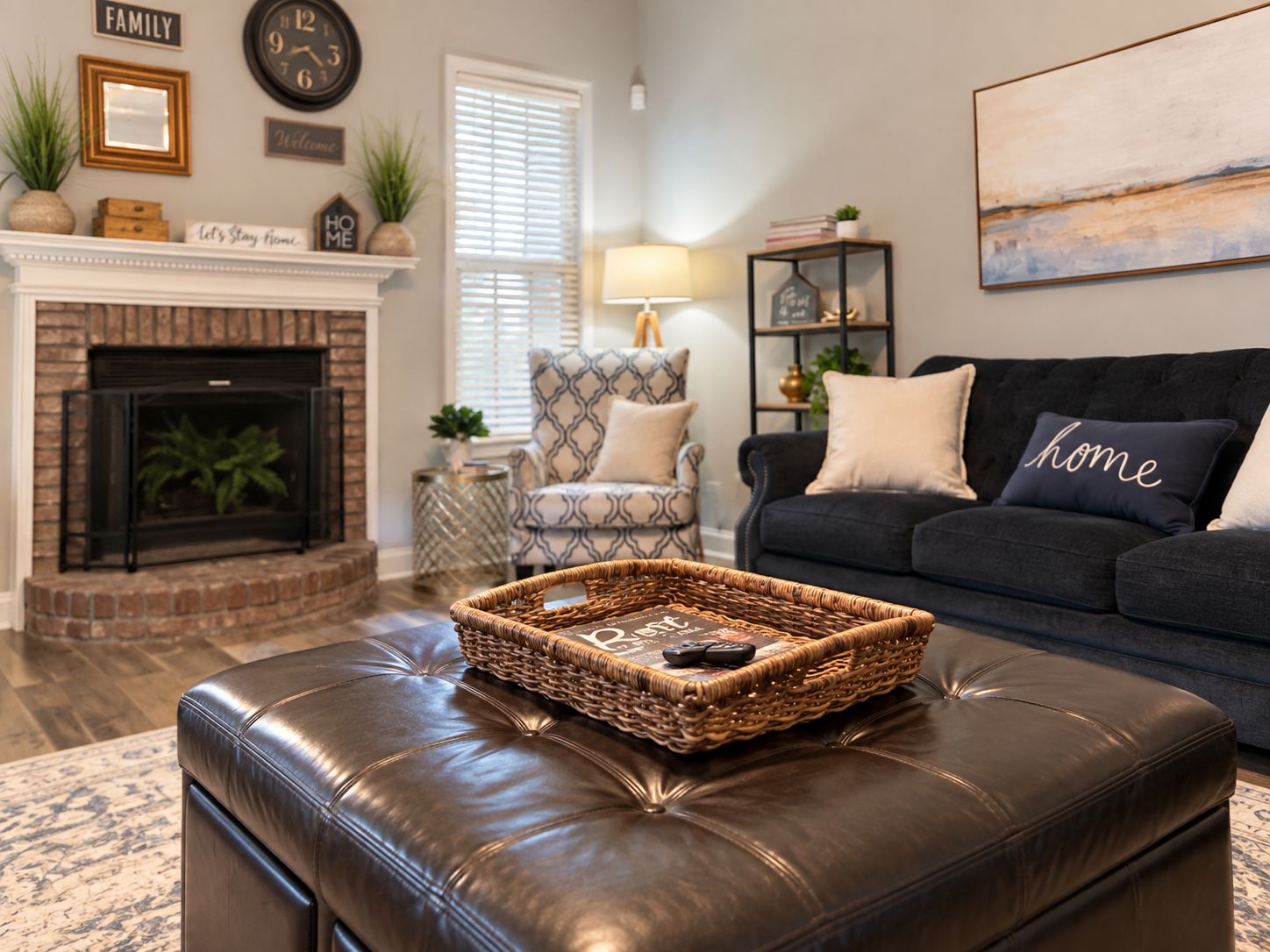 A living room with a fireplace, a patterned armchair, a dark sofa with a 'home' pillow, and a wicker tray on a leather ottoman.