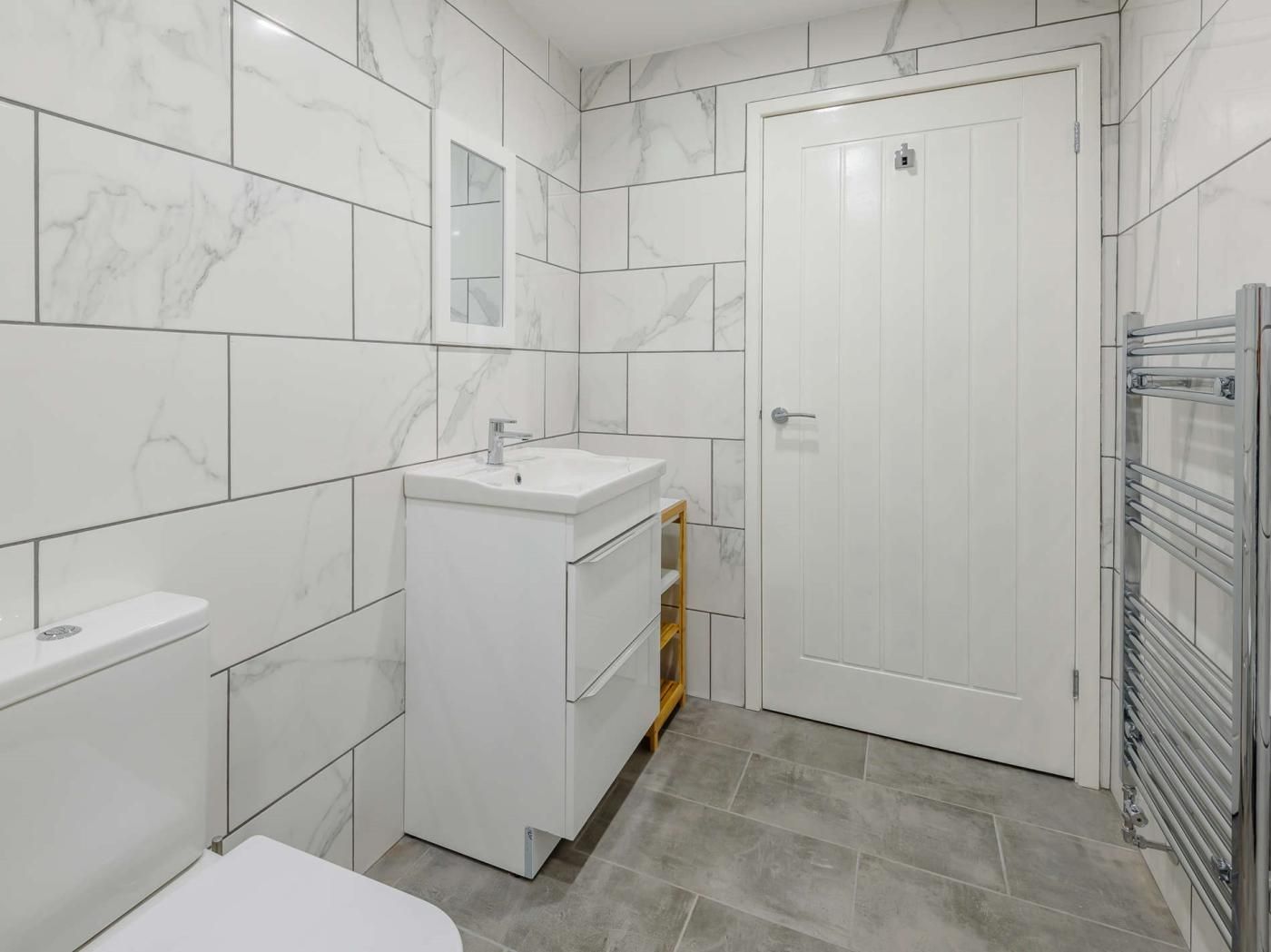 A bathroom with a white sink, mirror, and toilet, tiled in white with grey accents.