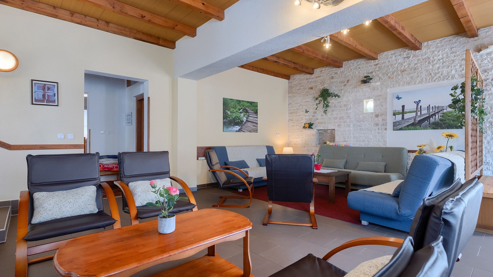 A cozy living room with a stone wall, wooden ceiling beams, and a variety of seating including sofas and chairs.