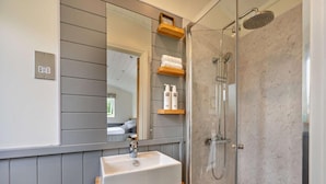A bathroom with a shower, a sink, a mirror, and a shelf with toiletries. - 1 Bed in Penrith (oc-85531) (Deepdale)