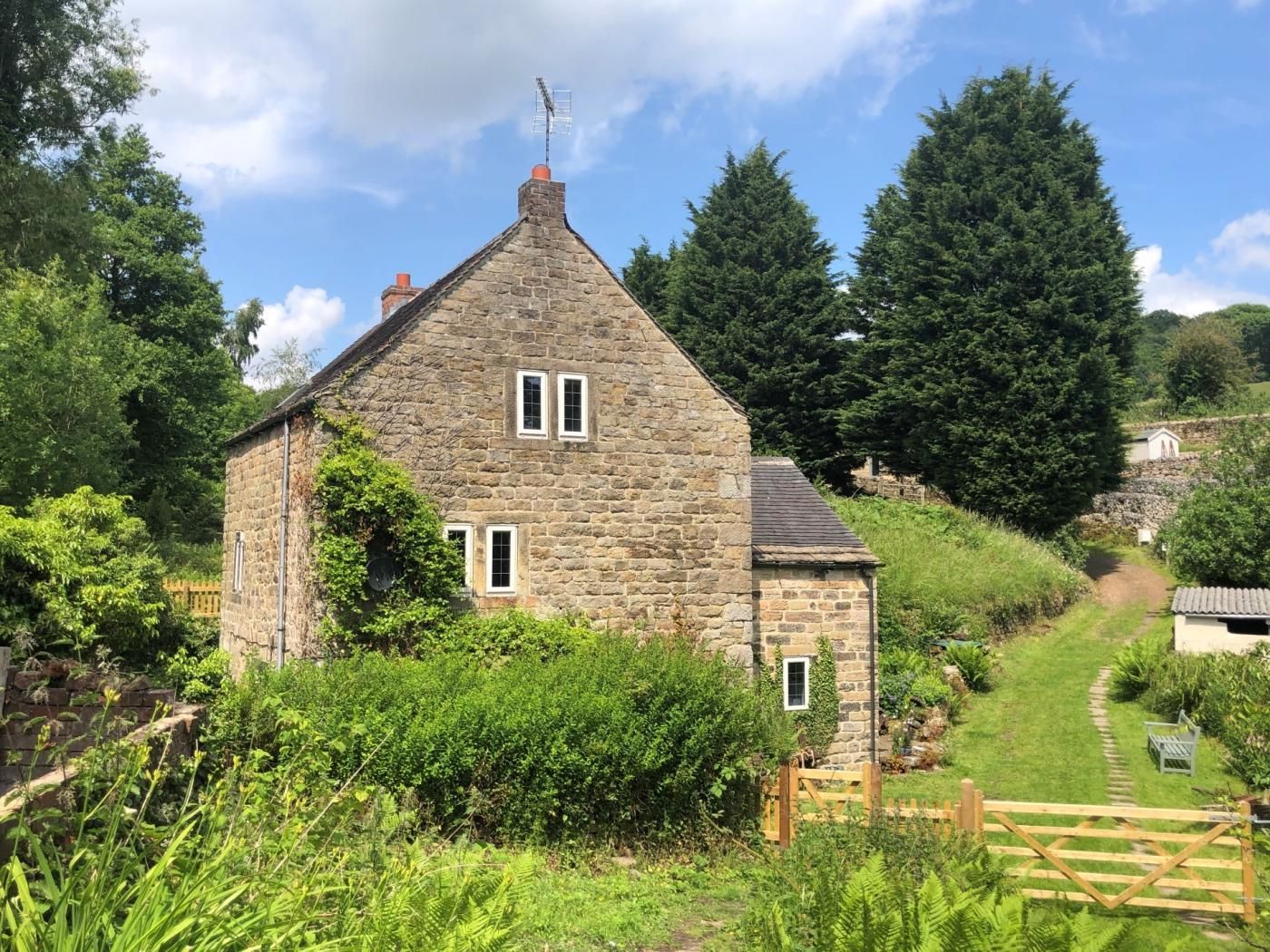 A stone house with a sloped roof, surrounded by greenery and a wooden gate.