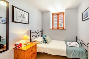 Ein Schlafzimmer mit Bett, Nachttisch, Lampe, Kommode, Fenster mit Jalousien und gerahmten Bildern an der Wand.