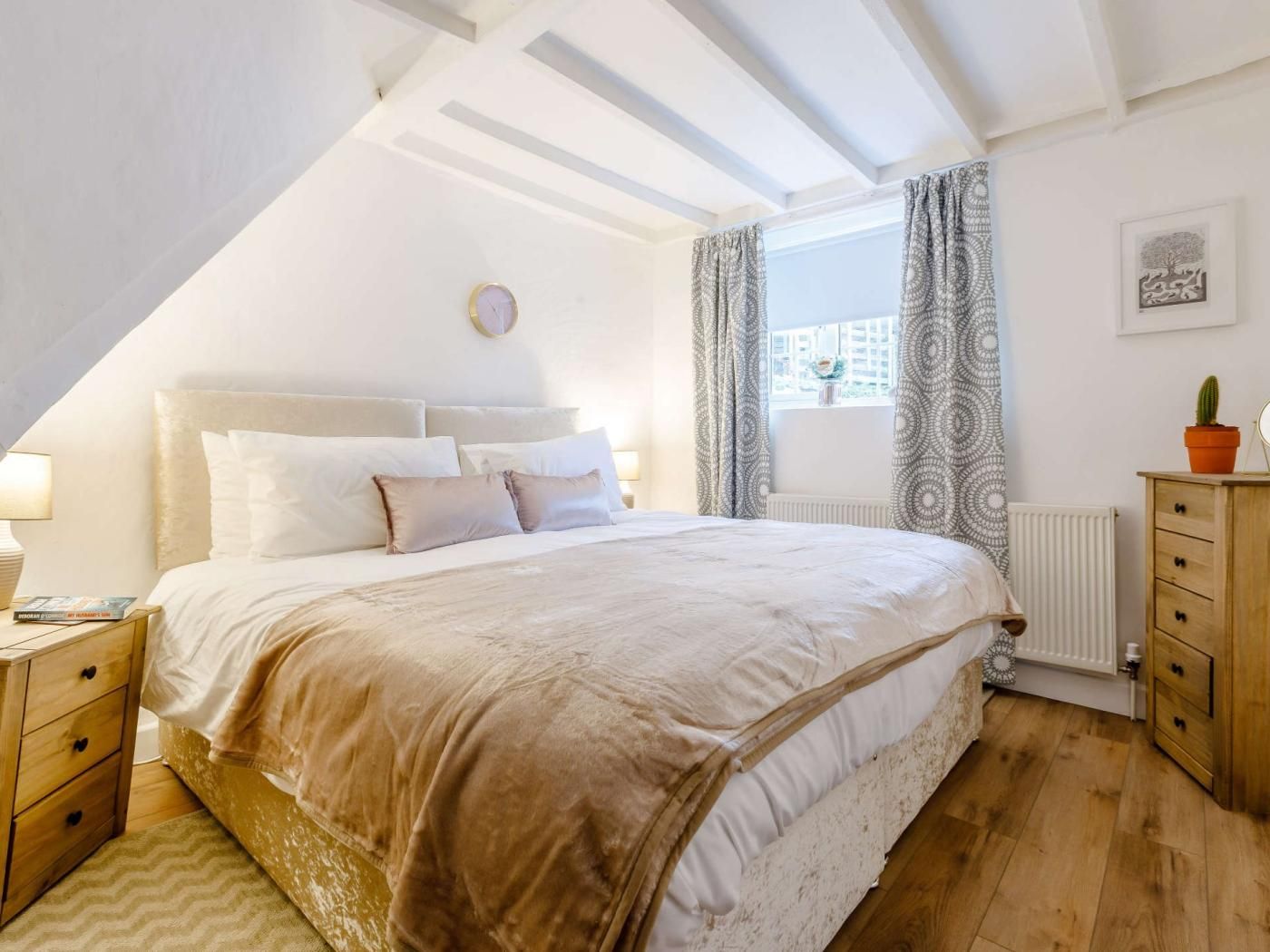 A bedroom with a large bed, a bedside table, a nightstand, a window with curtains, a radiator, and a wooden dresser.