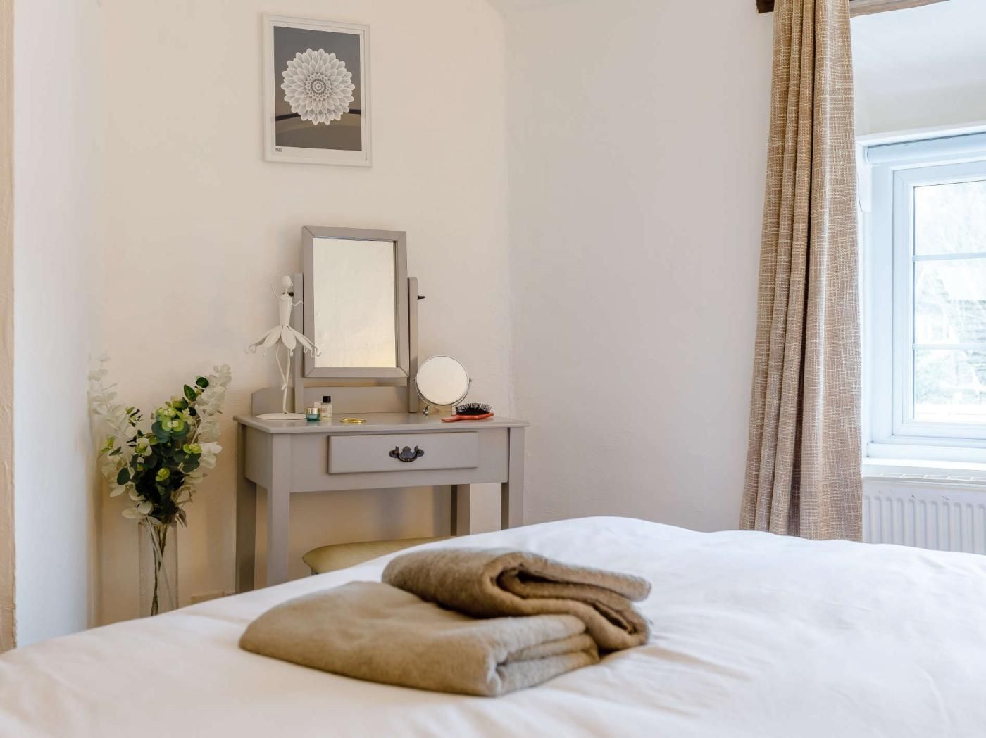 A hotel room with a bed, a bedside table with a mirror, a vase with flowers, and a window with curtains.