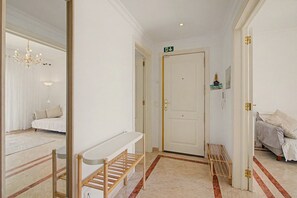 Ein Korridor mit einer weißen Tür, einer Bank, einem Kronleuchter und einem Bett im Hintergrund.