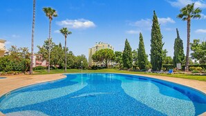 Uma piscina com água azul cristalina, cercada por palmeiras e ciprestes, com um edifício ao fundo.