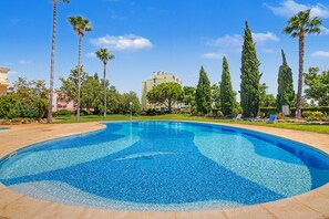 Ein Schwimmbecken mit klarem, blauem Wasser, umgeben von Palmen und Zypressen, mit einem Gebäude im Hintergrund.