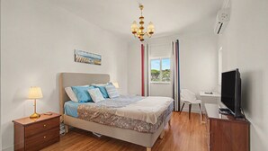 Quarto com cama, mesa de cabeceira, abajur, televisão e janela com cortinas.