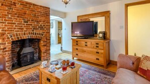 A living room with a brick fireplace, a television on a wooden stand, a leather sofa, and a coffee table with cups and pastries. - 3 Bed in Sandown (oc-85985) (Sandown)