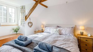 Une chambre avec un plafond en pente, un lit recouvert d’une couvre-lit à motifs, deux lampes de chevet, une table de chevet avec une horloge et une fenêtre avec des rideaux.
