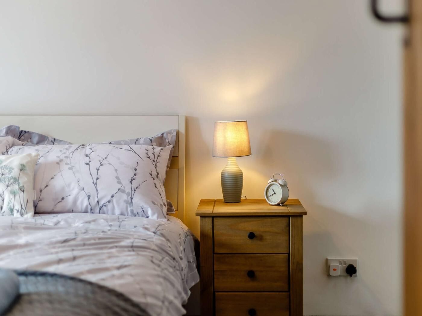 A neatly made bed with floral bedding, a bedside lamp, a small wooden nightstand with a clock, and a power outlet.