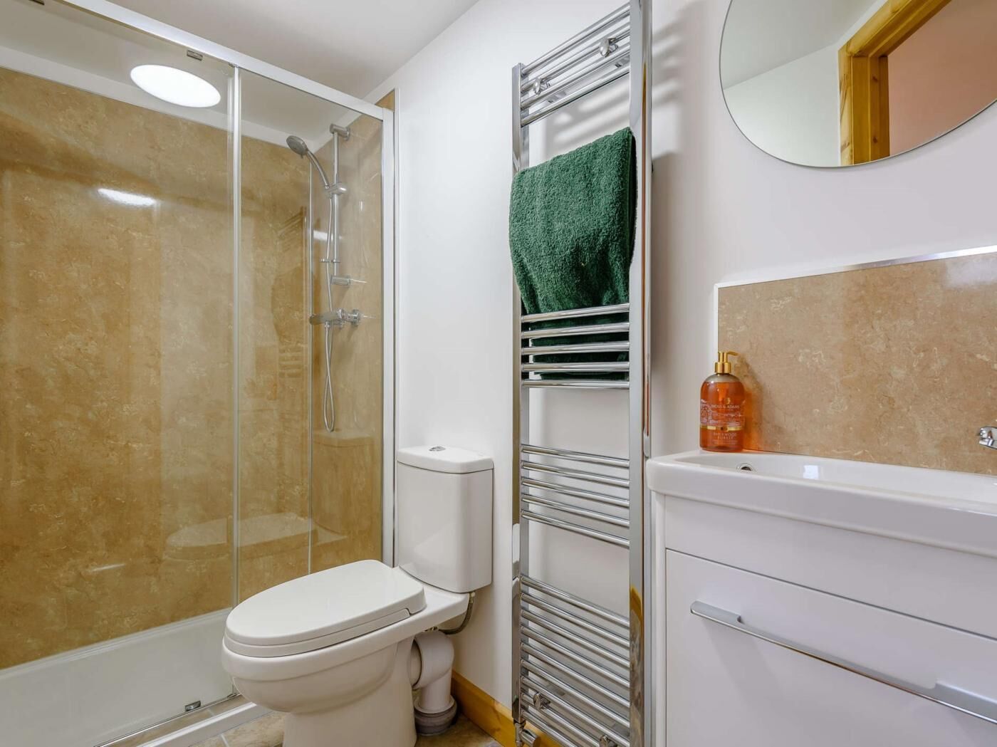 Un baño con cabina de ducha de vidrio, inodoro blanco, toallero con una toalla verde y lavabo con espejo arriba.