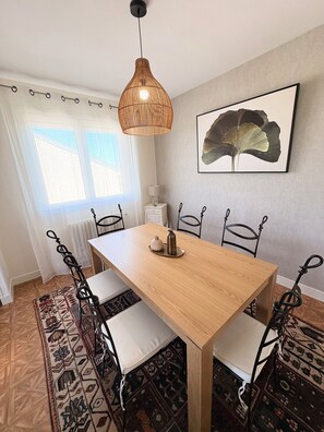 Een eetruimte met een houten tafel, zwarte metalen stoelen, een geweven hanglamp, een kamerkleed met een patroon en een kunstwerk in een lijst aan de muur.