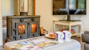 Ein gemütliches Wohnzimmer mit Kaminofen, Fernseher und Tisch mit Zeitschrift und Tasse.