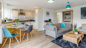 Una sala moderna con un sofá gris, sillas azules, una mesa de centro y una cocina con armarios blancos y lámparas colgantes azules.