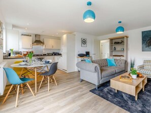 A modern living room with a grey sofa, blue chairs, a coffee table, and a kitchen area with white cabinets and blue pendant lights. - 2 Bed in Y Felinheli (oc-86260) (Y Felinheli)
