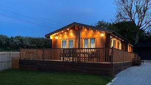 A wooden cabin with a deck, a covered patio, and a gravel driveway.