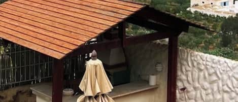 Uma área de estar coberta ao ar livre com mesa e cadeiras, uma pequena cozinha e vista para um vinhedo.