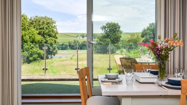 Ein Essbereich mit Tisch für zwei Personen, Blick auf eine grüne Landschaft und ein Fenster mit Vorhängen.
