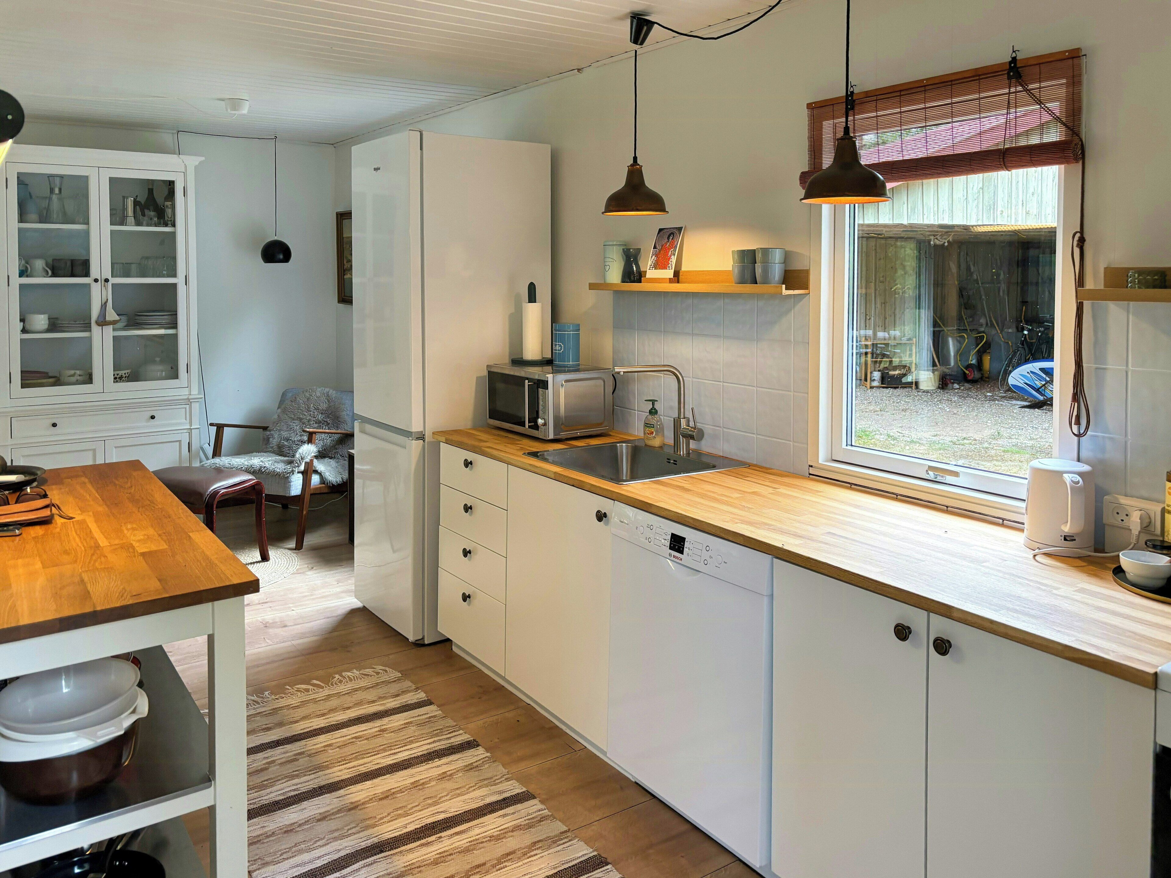 A modern kitchen with white cabinetry, a wooden countertop, a built-in dishwasher, a microwave, and a window with a view of the outside.