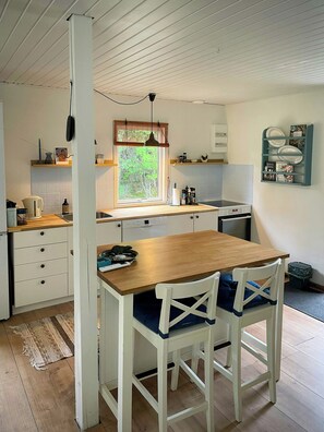 Una cocina con una mesa de madera, sillas blancas, un ventanal con vistas a árboles y un escurreplatos fijado a la pared.