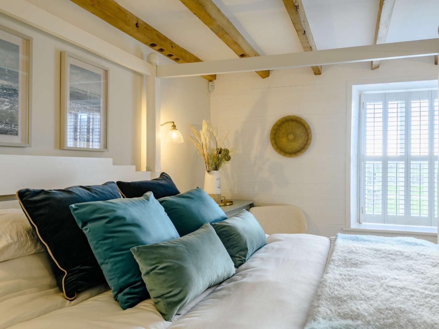 A bedroom with a bed, a nightstand, a wall-mounted lamp, a window with blinds, and a decorative plate.