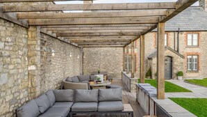 A covered outdoor seating area with stone walls, a wooden pergola, and a view of a stone building. - 8 Bed in Bridgend (oc-86493) (Southerndown)