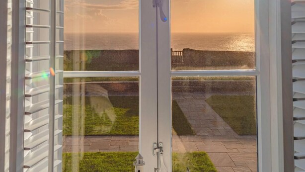 A view through a glass door to a patio with a stone pathway, a grassy area, and a body of water in the background. - 8 Bed in Bridgend (oc-86493) (Southerndown)