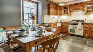 A kitchen with wooden cabinets, a dining table set with plates and glasses, and a window with a view. - 3 Bed in Tenby (oc-86625) (Tenby)
