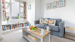 A modern living room with a grey sofa, a coffee table with fruits and a mug, a bookshelf, and a window with curtains. - 3 Bed in Bude (oc-86665) (Bude)