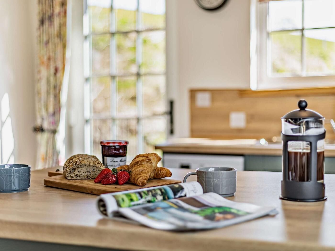 Une table en bois dressée pour le petit-déjeuner, avec des croissants, des fraises, de la confiture, une cafetière à piston et un journal.