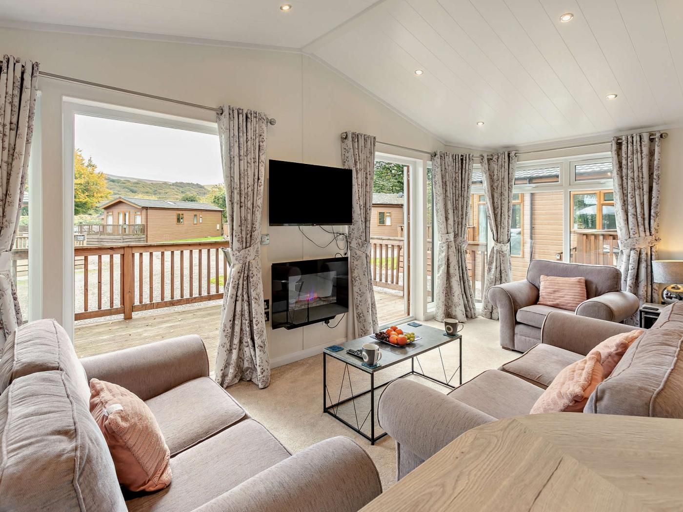 A spacious living room with a flat-screen TV mounted on the wall, a comfortable sofa set, a coffee table, and a balcony with a wooden railing.