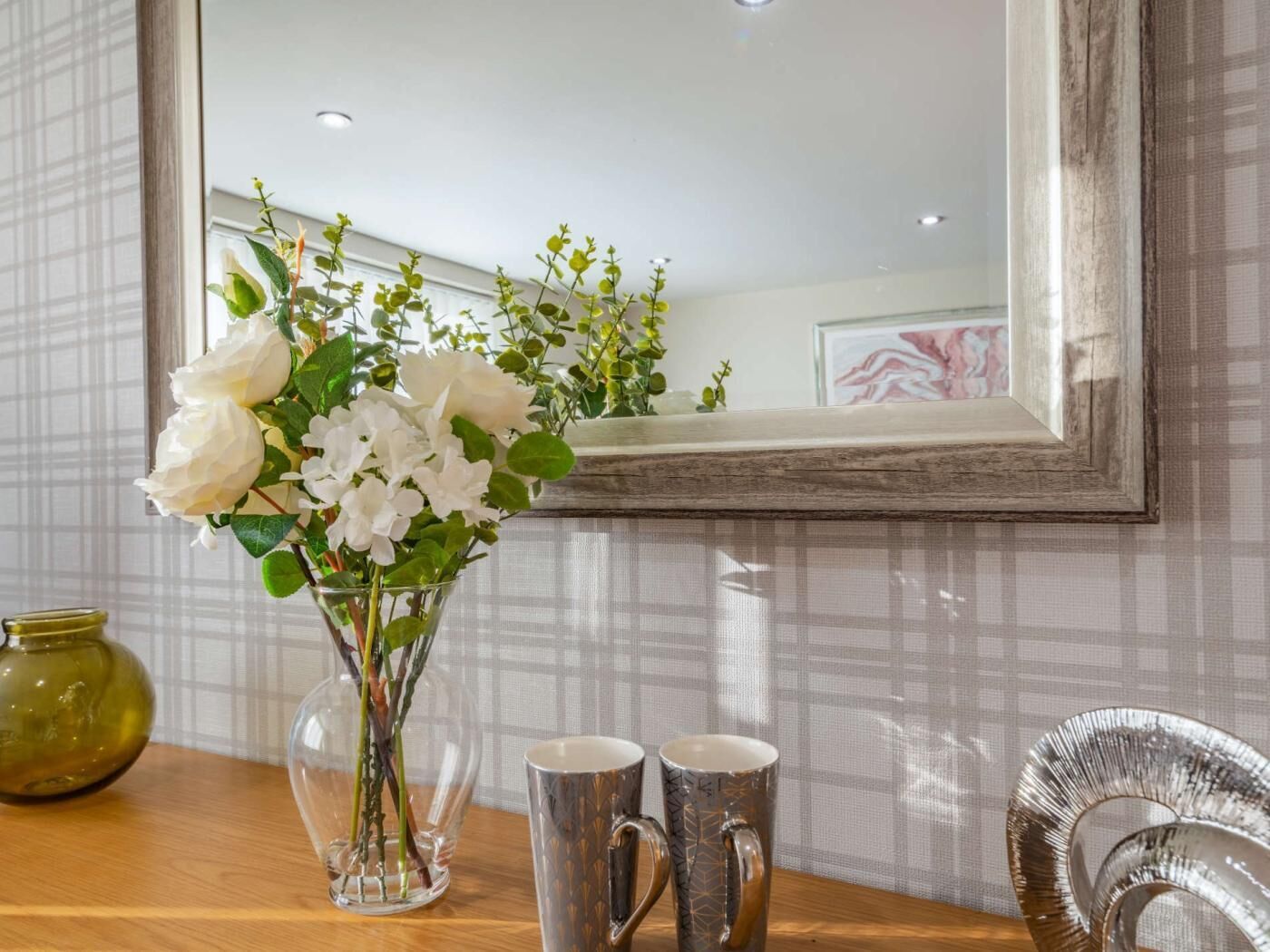 Una habitación con un espejo, un florero con flores blancas, dos tazas metálicas y un florero verde sobre una mesa de madera.