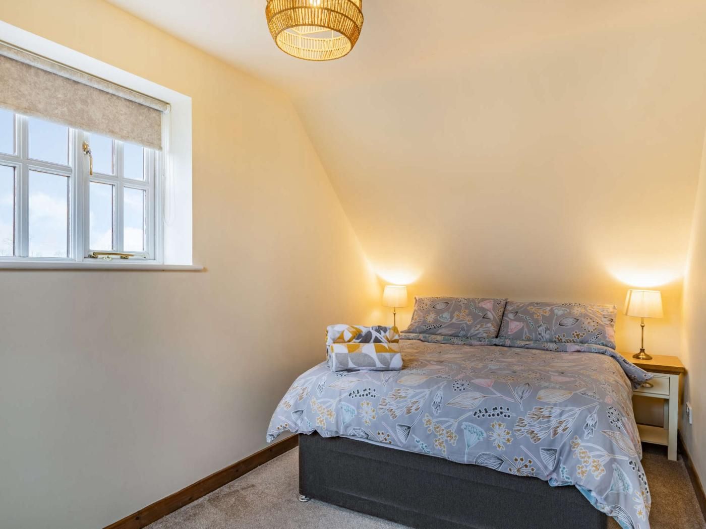 A bedroom with a bed, bedside lamps, a window with a roller blind, and a hanging light fixture.