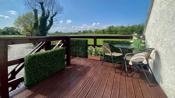 A wooden deck with a table and chairs, a hedge, and a view of a green field under a blue sky. - 1 Bed in Hawarden (oc-86711) (Dobshill)