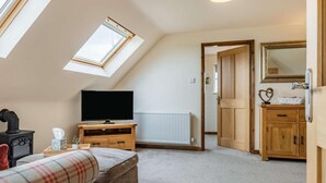 A room with a sloped ceiling, a television on a wooden stand, a radiator, a wooden cabinet, and a door leading to another room. - 1 Bed in Hawarden (oc-86711) (Dobshill)