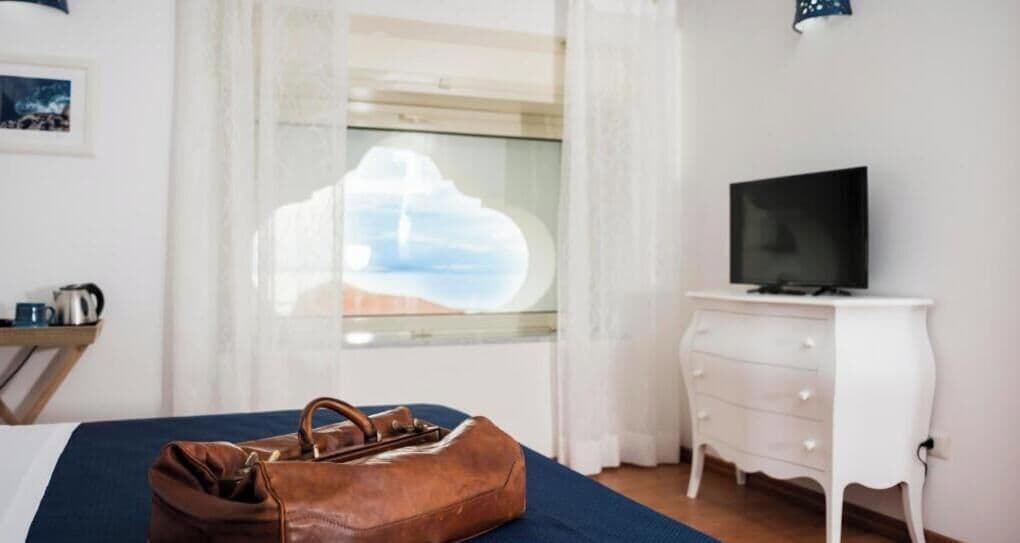 Hotelkamer met een bed, een televisie op een dressoir, een raam met gordijnen en een bruine tas op het bed.