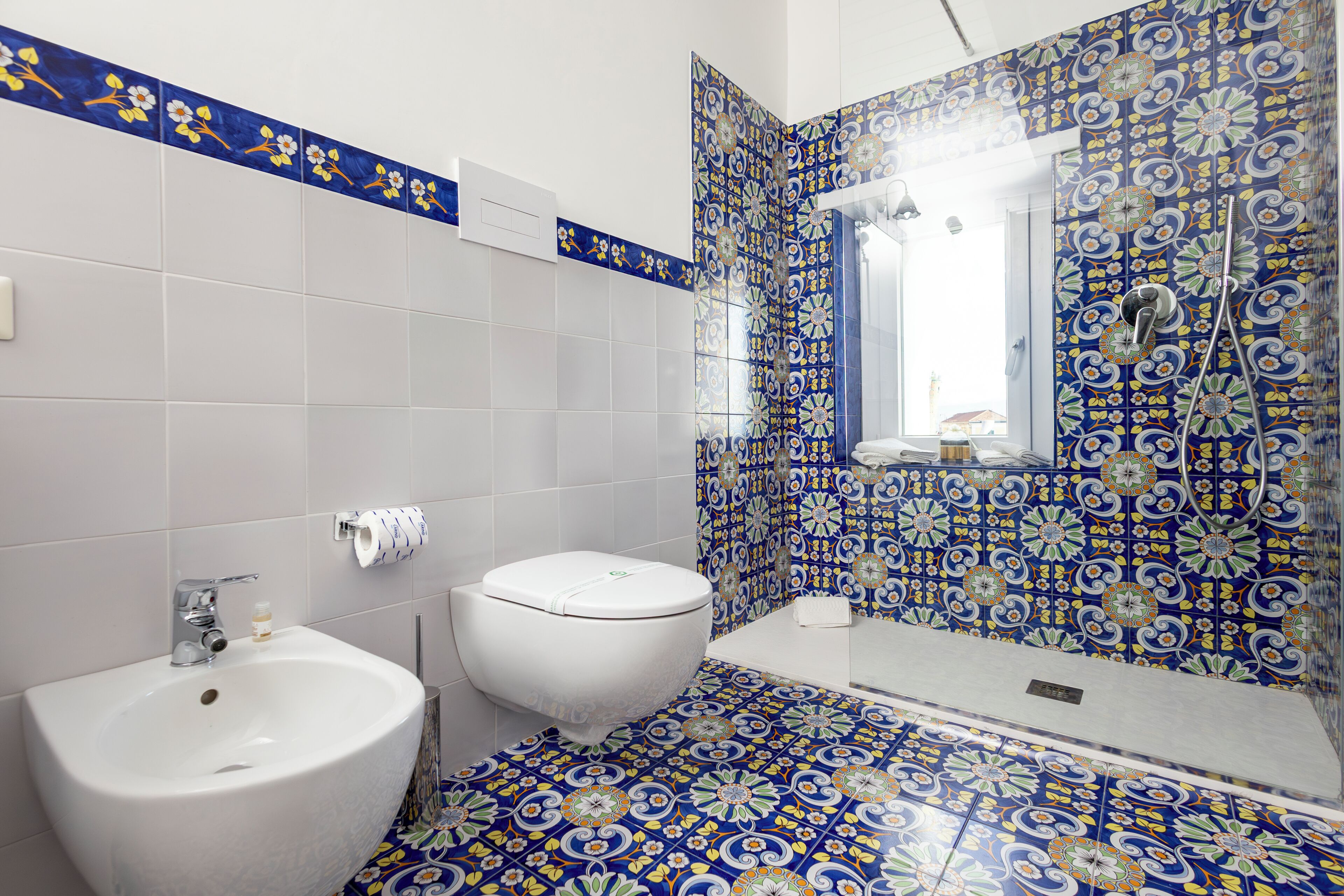 Baño con lavamanos, inodoro y zona de ducha, con azulejos blancos y azules estampados.