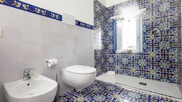Baño con lavamanos, inodoro y zona de ducha, con azulejos blancos y azules estampados.