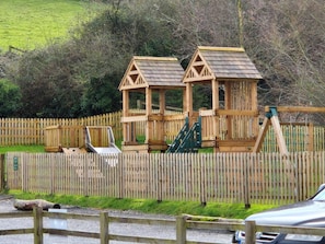 A wooden play structure with a slide and climbing area, surrounded by a wooden fence. - 3 Bed in Minehead (oc-87069) (Withycombe)