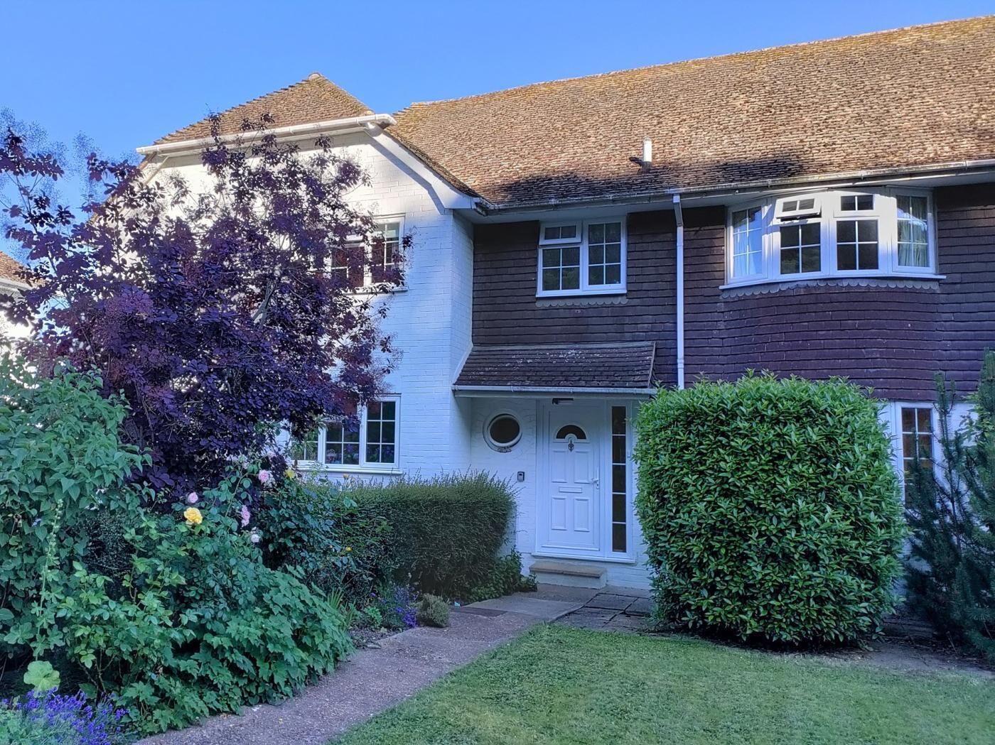 A two-story house with a white front door, a small front porch, and a well-maintained garden with various plants and a hedge.