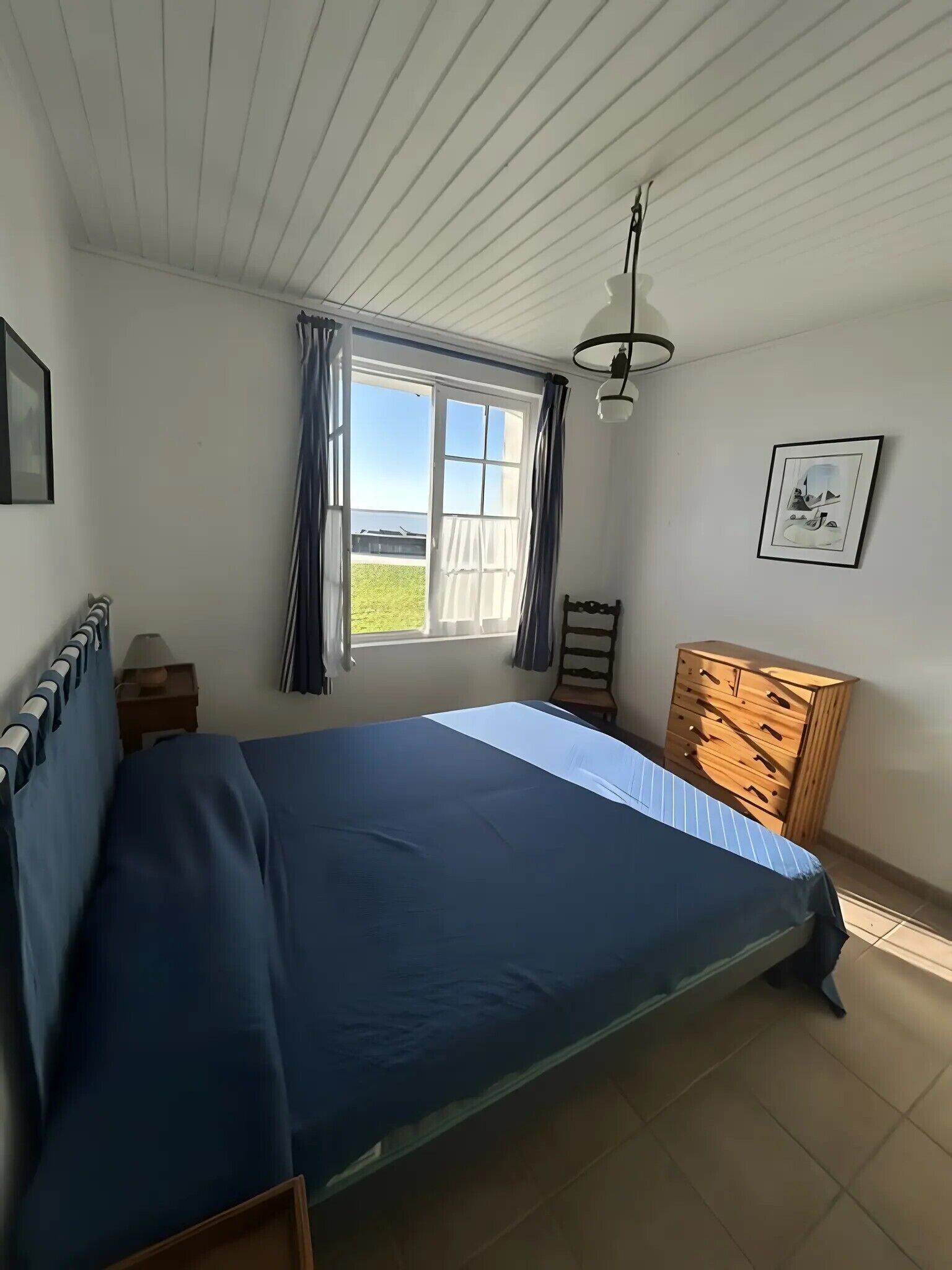 A bedroom with a bed, a wooden dresser, a window with curtains, a framed picture on the wall, and a ceiling light.