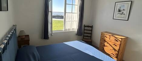 A bedroom with a bed, a wooden dresser, a window with curtains, a framed picture on the wall, and a ceiling light.
