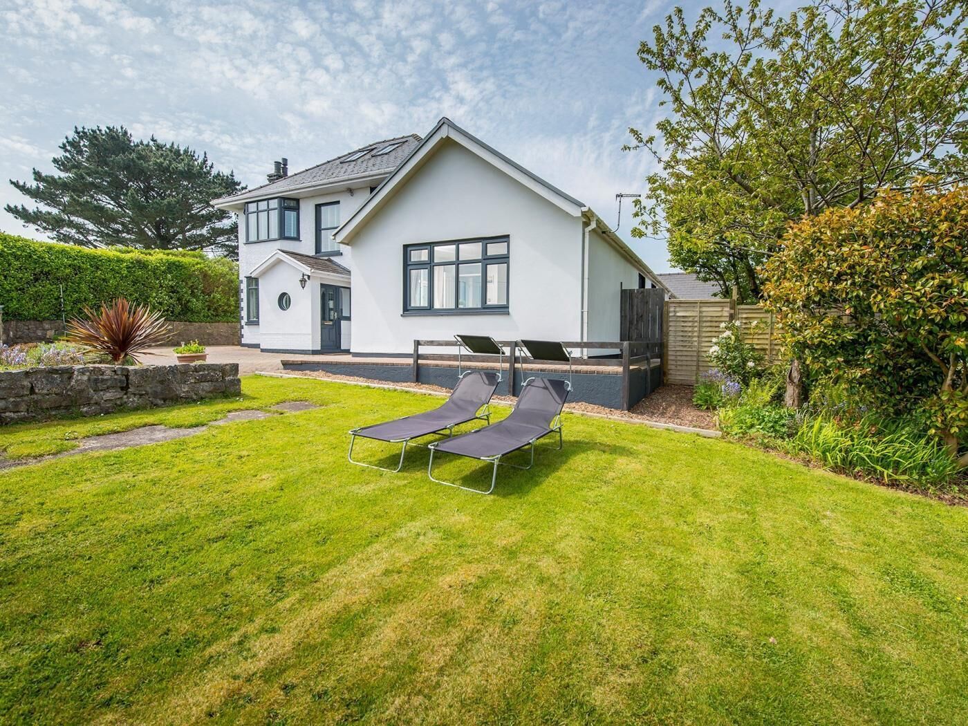 A modern two-story house with a well-maintained lawn, two sun loungers, and a wooden fence.