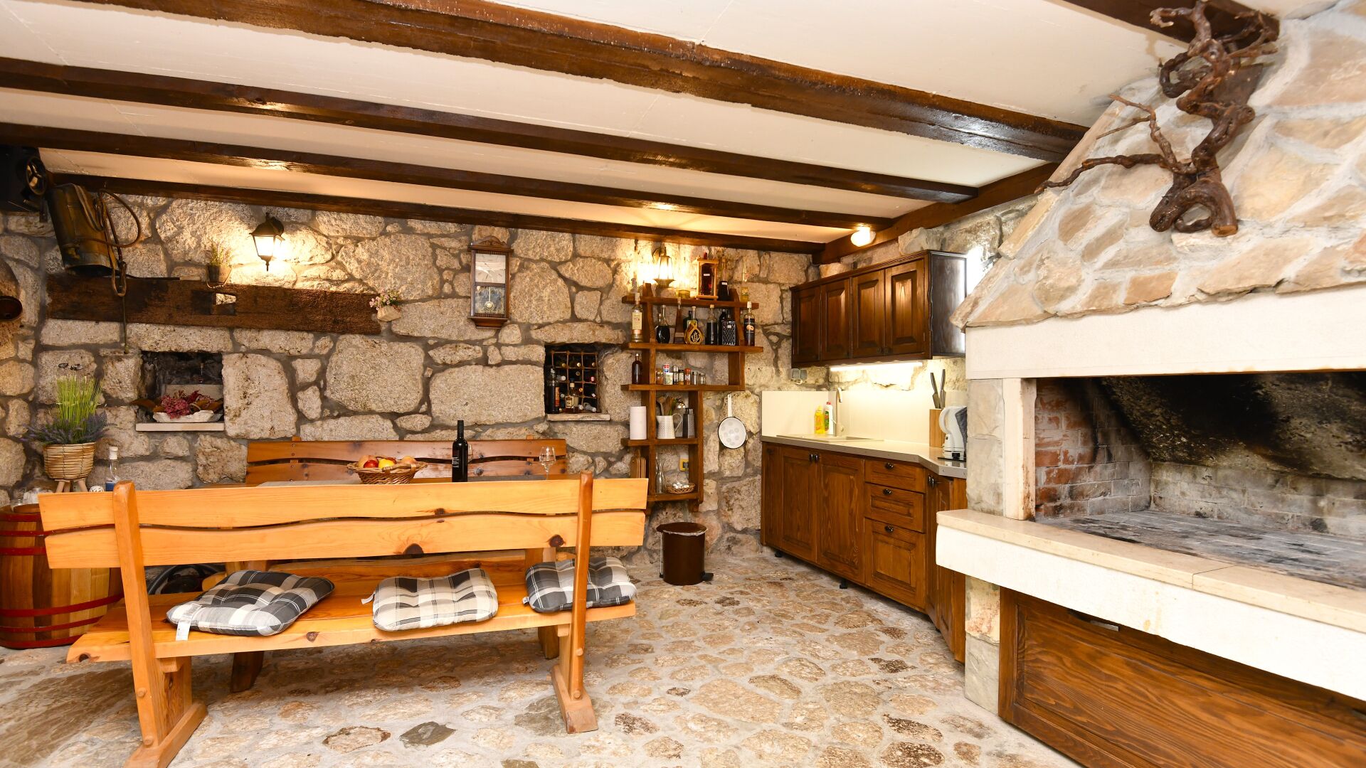 A rustic kitchen with stone walls, wooden beams, a stone fireplace, and a wooden bench with cushions.