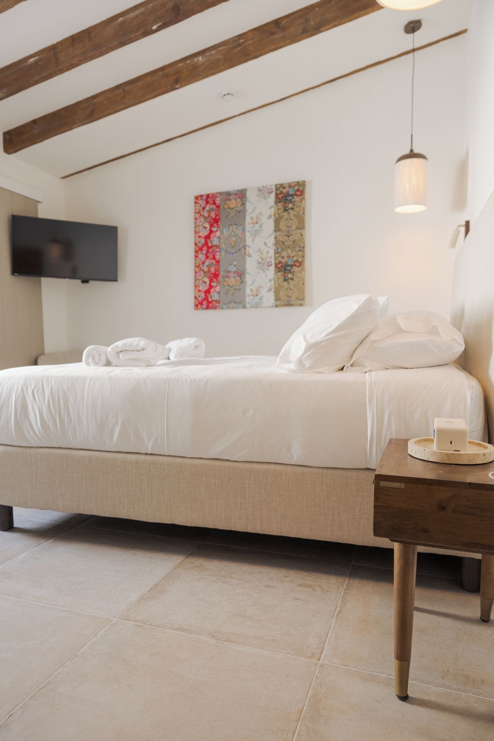 Un dormitorio con cama, almohadas, mesita de noche, televisor de pantalla plana y una obra de arte colgada en la pared.